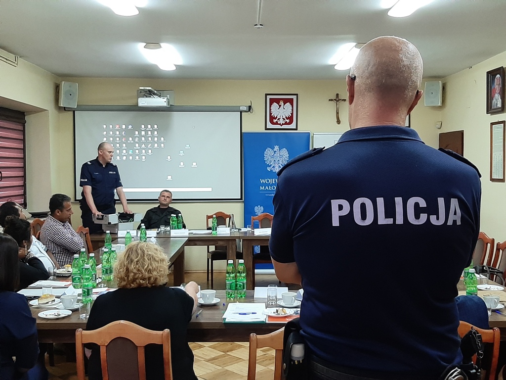 Szkolenie prowadzone przez policjantów, na pierwszym planie widać tył munduru policyjnego,szkolenie odbywa się w sali gdzie stoły ułożone są w kwadrat